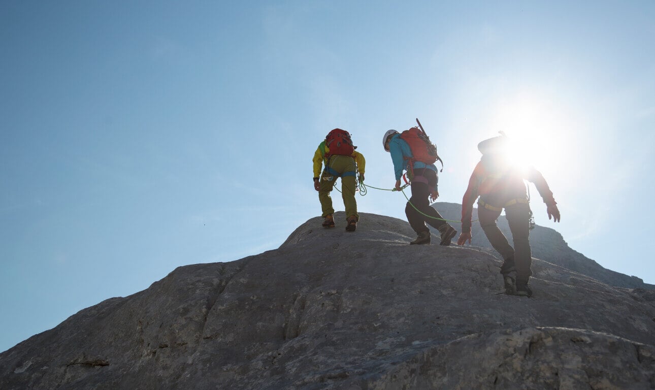 Three climbers ascending a rocky slope under clear skies, roped together for safety, symbolizing teamwork, resilience, and shared goals